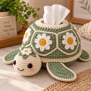 Peut inclure: Une housse de boîte à mouchoirs en crochet en forme de tortue avec une carapace vert sauge ornée de motifs de marguerites blanches et jaunes. La tête est crème avec des yeux noirs, des joues roses et une petite marguerite. Un mouchoir blanc dépasse.