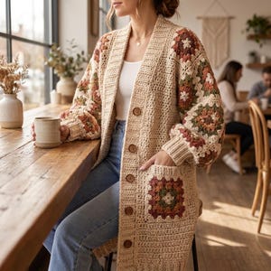 Op de afbeelding: Een beige gehaakt vest met lange mouwen en een granny square-patroon in bruin-, groen- en crèmetinten. Het kledingstuk heeft een V-hals, een knoopsluiting aan de voorzijde met vijf bruine knopen en twee opgestikte zakken met bijpassende motieven.