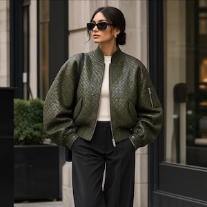 May include: An olive green bomber jacket with a woven, quilted texture, featuring a ribbed collar, cuffs, and hem, a front zipper closure, and side pockets. The jacket is worn over a white top and paired with black trousers and black sunglasses.