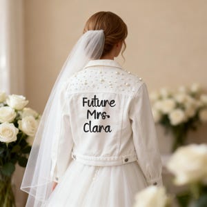 Puede incluir: Una chaqueta vaquera blanca con cuello, mangas largas y cierre de botones. La parte trasera está adornada con perlas blancas dispersas en el canesú y el texto bordado en negro Future Mrs. Clara centrado en el panel posterior.