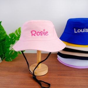 Puede incluir: Un sombrero de pescador de color rosa claro con el nombre Rosie bordado en hilo rosa oscuro en la parte delantera. El sombrero cuenta con una correa negra para la barbilla y se exhibe en un soporte de madera. Se ve una pila de otros sombreros.