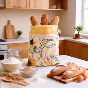 Puede incluir: Una bolsa de pan de tela color crema con un estampado de girasoles, trigo y panales, con el texto "Organic Bread" y el nombre "Oliver" bordado en azul. La bolsa está llena con tres barras de pan y tiene un forro interior de color amarillo.