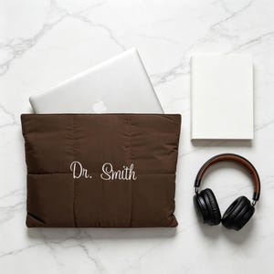 May include: A dark brown quilted laptop sleeve featuring the white embroidered text Dr. Smith, shown with a silver laptop partially inserted, a white rectangular notebook, and black over-ear headphones with brown accents on a marble surface.