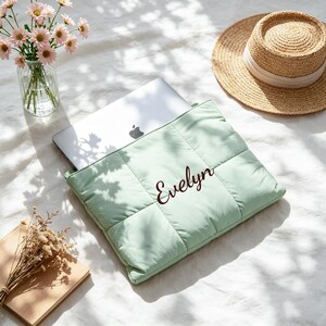 May include: A light green quilted laptop sleeve featuring the name Evelyn embroidered in dark brown script. The rectangular padded case holds a silver laptop, showcasing a soft, stitched grid pattern across its surface for protection.