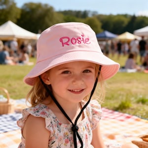 Puede incluir: Un sombrero de pescador de color rosa claro con el nombre Rosie bordado en una fuente cursiva de color rosa oscuro en la parte delantera. El sombrero incluye una correa negra para la barbilla con un cierre ajustable para mayor sujeción.