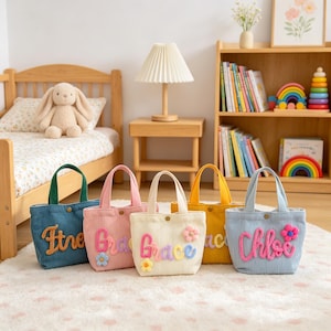 Puede incluir: Cinco bolsos pequeños de pana en azul, rosa, crema, amarillo y azul claro. Cada bolso presenta un nombre personalizado en letras coloridas, texturizadas y en relieve, con pequeñas flores de tela decorativas en la parte frontal.