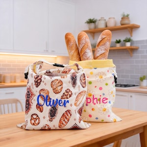 May include: Two fabric bread bags with roll-top closures. The left bag is cream-colored with a pattern of various breads and the name Oliver in blue. The right bag is cream-colored with a daisy and polka dot pattern and the name Sophie in pink.