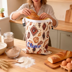 May include: A cream-colored fabric bread bag featuring a repeating pattern of various baked breads and geometric shapes, with the name Oliver embroidered in blue script. The bag has a white top trim with a black plastic buckle closure.