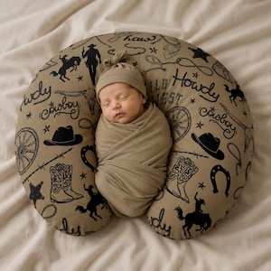 Puede incluir: Un bebé recién nacido envuelto en una muselina beige y un gorro a juego, descansando sobre una almohada de lactancia en forma de herradura. La almohada es beige con ilustraciones negras con temática de vaqueros y las palabras "Howdy Cowboy" y "Wild West".
