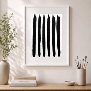 Op de afbeelding: Een ingelijste wandkunstprint met zes verticale zwarte penseelstreken op een witte achtergrond. Het kunstwerk zit in een dunne witte lijst en toont een abstract ontwerp met een hoog contrast en textuurrijke verfstreken.