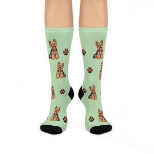May include: A pair of light green crew socks featuring a repeating pattern of small Yorkshire Terrier dogs and dark brown paw prints. The socks have solid black heels and toe sections, providing a distinct contrast to the light green fabric.