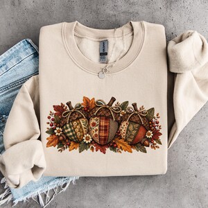 Op de afbeelding: Een beige sweater met ronde hals voorzien van een afbeelding van drie grote eikels versierd met diverse herfstachtige stofpatronen, omringd door herfstbladeren, kleine witte bloemen en rode bessen. Het Gildan-merklabel is zichtbaar bij de hals.