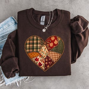 Pode incluir: Uma camisola de gola redonda castanho-escura com um grande gráfico em forma de coração composto por vários padrões de patchwork, incluindo xadrez, folhas de outono, estampados florais e vichy, acentuado com um pequeno botão e um laço.