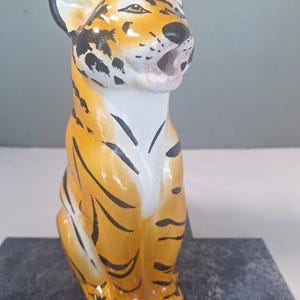 May include: A ceramic tiger figurine in a seated position with an open mouth. The tiger features a bright orange coat with black stripes, a white chest and muzzle, and black-tipped ears. The surface has a glossy finish and detailed facial features.