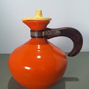 May include: An orange ceramic jug with a yellow lid and a dark wood handle. The jug has a rounded body and a flared top. A metal band secures the handle to the jug.