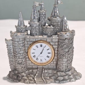 Pode incluir: Um relógio de metal prateado em forma de castelo com um mostrador circular central, aro dourado, fundo branco, algarismos romanos pretos e ponteiros pretos. A estrutura inclui várias torres, texturas de tijolos e plantas trepadeiras.