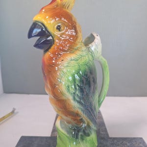 May include: A ceramic pitcher shaped like a parrot with a glossy finish. The bird features a vibrant orange and yellow head with a dark blue beak, transitioning into green and dark blue feathers on the body. A handle is attached to the back.