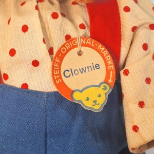 May include: A vintage Steiff Clownie doll, featuring a cream-colored shirt with red polka dots, blue overalls, and a red felt bib. The doll has a tag that reads "Clownie" with the Steiff logo.