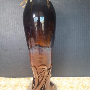 May include: A tall, cylindrical ceramic vase featuring a glossy dark brown glaze at the top that transitions into a lighter, earthy terracotta tone at the base. The lower section is adorned with intricate, raised, swirling clay relief patterns.