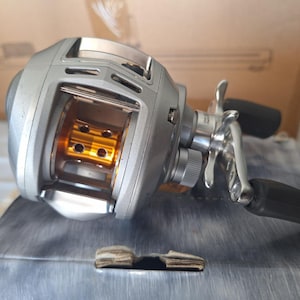 Può includere: Un mulinello da pesca baitcasting color argento con una bobina in metallo dorato dotata di perforazioni circolari. Il mulinello include una manovella argentata con due impugnature in gomma nera, una manopola di regolazione e una frizione.
