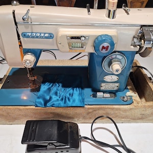 May include: A vintage Morse Zig Zag sewing machine featuring a two-tone cream and blue metal body. The machine includes a stitch length dial, a stitch pattern selector, a needle assembly, and a black foot pedal connected by a power cord.