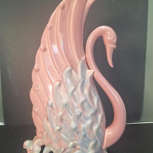 Puede incluir: Una figura de cisne de cerámica en tonos rosa, azul y blanco. El cisne tiene un cuello largo y curvado y alas detalladas. La base presenta un diseño texturizado con agujeros. Un objeto decorativo.