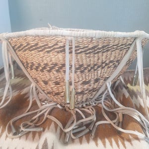 May include: A woven basket with a conical shape featuring alternating light and dark brown geometric patterns. The rim is wrapped in light-colored leather strips that extend downward into long fringe, each tipped with a polished metallic cone.