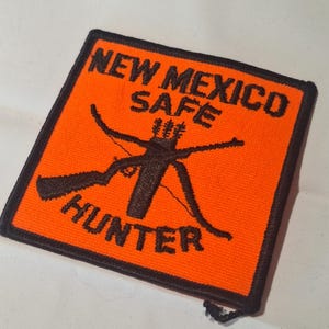 May include: A square embroidered patch with a bright orange background and a black border. The text NEW MEXICO SAFE HUNTER is stitched in black. A black graphic of a crossed rifle and bow with a quiver of arrows is centred on the patch.