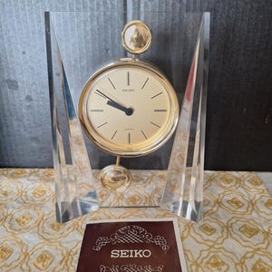 Puede incluir: Un reloj de sobremesa de cuarzo Seiko con esfera circular dorada y manecillas negras, suspendido en un marco de acrílico transparente. Un péndulo dorado cuelga bajo la esfera. Un manual de instrucciones marrón se sitúa delante.