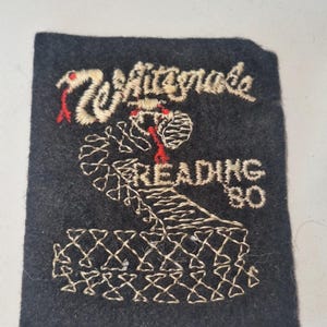 May include: A rectangular black fabric patch featuring white embroidered text that reads Whitesnake and READING 80. The design includes a central embroidered cobra snake in white thread with red accents for the tongue and eyes, set against a dark background.
