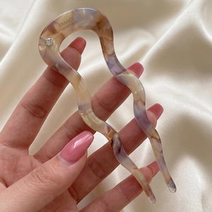 Puede incluir: Una horquilla para el cabello ondulada de dos puntas en tonos beige, morado y crema. El accesorio para el cabello tiene un pequeño pedrería transparente y el texto "FRANCE Handmade". La horquilla se sostiene en una mano.