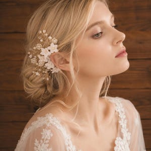 May include: A delicate bridal hair comb featuring white fabric flowers, small pearls, and gold-colored wire. The floral hair accessory is designed to be worn in the hair for weddings or special occasions. The comb is shown on a person with blonde hair.