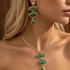 May include: Gold-toned jewelry set featuring a necklace and earrings. The necklace has a delicate chain with a pendant of three green, marbled-effect stones. The earrings have a similar design, with three green stones suspended from a gold disc.