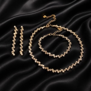 Könnte beinhalten: Ein goldfarbenes Schmuckset auf schwarzem Satinhintergrund. Das Set umfasst eine Halskette, ein Armband und Ohrringe, alle mit einem Zickzackmuster aus kleinen, funkelnden Steinen. Die Ohrringe sind lang und linear.
