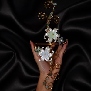 Può includere: Un accessorio per capelli floreale color oro con fiori bianchi e azzurri e accenti verdi. Il design presenta un delicato lavoro di fili e piccole pietre decorative, creando un look elegante ispirato alla natura.