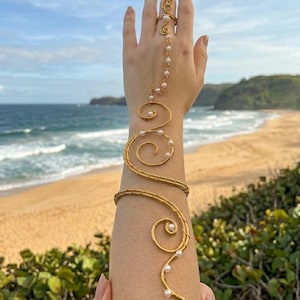 May include: A gold-toned wire arm cuff and ring set featuring a continuous spiral design that wraps around the forearm and connects to a finger ring via a delicate chain. The metal wire is accented with small white pearls throughout the structure.