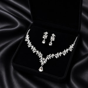 Può includere: Un set di collana e orecchini argentati, presentato in un cofanetto di velluto nero. I gioielli presentano un design floreale con cristalli chiari e scintillanti. La collana ha un pendente a goccia. Il set è esposto su uno sfondo di raso nero.