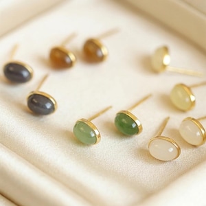 Op de afbeelding: Een verzameling oorstekers met ovale edelstenen in verschillende kleuren, waaronder grijs, bruin, groen en wit. Elke oorbel heeft een goudkleurige bezel en pin. De oorbellen worden gepresenteerd op een wit, getextureerd oppervlak.