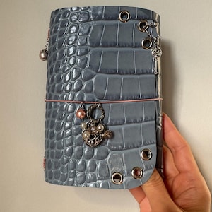 May include: A blue-grey journal cover with a textured crocodile skin pattern, featuring silver-toned metal eyelets along the edges. The cover is secured with a thin pink elastic band adorned with a dangling heart charm and several small beads.