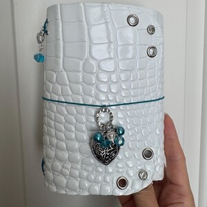 May include: A white journal cover with a textured crocodile skin pattern, featuring silver metal eyelets and a teal elastic band. A silver heart-shaped charm with intricate cutouts and teal beads hangs from the center of the elastic band.