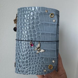 May include: A light blue notebook cover with a textured crocodile skin pattern, featuring gold-toned eyelets, small embedded crystals, and dangling charms including a pink ladybug and a silver butterfly with clear stones attached to a black elastic band.