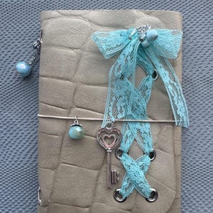 May include: A beige journal with a textured crocodile-skin pattern, featuring a light blue lace ribbon threaded through metal eyelets, a silver-toned heart-shaped key charm, and two light blue spherical beads attached to the cover.