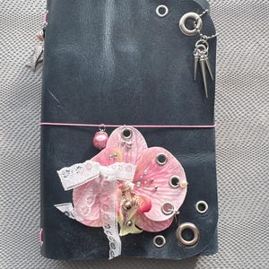 May include: A black journal cover featuring silver metal eyelets, a pink elastic band, and decorative charms including silver spikes, a pink pearl, and a pink orchid flower embellishment with a white lace ribbon tied around the center.
