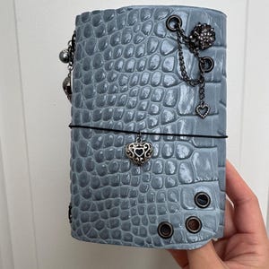 May include: A light blue journal cover with a textured crocodile skin pattern. The cover features dark metal grommets, a black elastic band, and silver-toned charms including a heart, a flower, and a small sphere attached by thin chains.