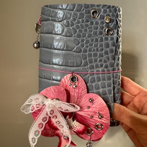 May include: A grey, crocodile-textured leather notebook cover featuring a pink elastic band, silver metal grommets, and a decorative pink orchid flower adorned with a white lace ribbon, small silver studs, and a dangling silver wing charm.