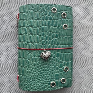 May include: A teal green journal cover with a textured crocodile skin pattern. The cover features silver metal eyelets along the right edge and is secured by a thin red elastic cord adorned with a silver filigree heart-shaped charm.