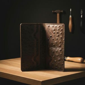 Può includere: Un portafoglio bifold marrone caratterizzato da una texture in pelle di struzzo con molteplici scomparti interni per carte e un orientamento verticale. Il portafoglio presenta cuciture visibili lungo i bordi e una distintiva trama a rilievo.