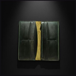 Può includere: Un portafoglio lungo in pelle verde scuro dal design a libro, dotato di molteplici fessure verticali per carte su entrambi i lati e un dorso interno in camoscio giallo. Il portafoglio è rifinito con cuciture visibili lungo i bordi e le tasche.