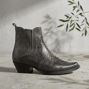 May include: A grey ankle boot featuring a pointed toe, a stacked block heel, and elastic side panels. The boot is constructed with a combination of smooth leather and a textured, snakeskin-patterned leather overlay with decorative stitching.
