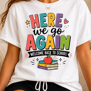 May include: A white t-shirt featuring a colorful graphic with the text "HERE we go AGAIN" in large, multi-colored letters. Below the text, a black banner reads "WELCOME BACK TO SCHOOL!" with a red apple resting on a stack of three books.